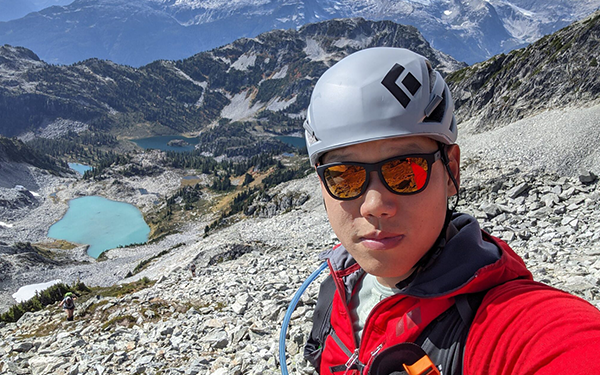 Jimmy selfie in helmet and sunglasses with a view of mountains and lakes